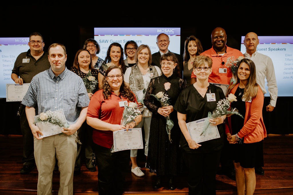 Mosaic Life Care School at Work, 2019 Graduates.  15 people are standing clustered together holding flowers and certificates. They are standing in front of three projection screens showing "SAW Guest Speakers" at their graduation ceremony.