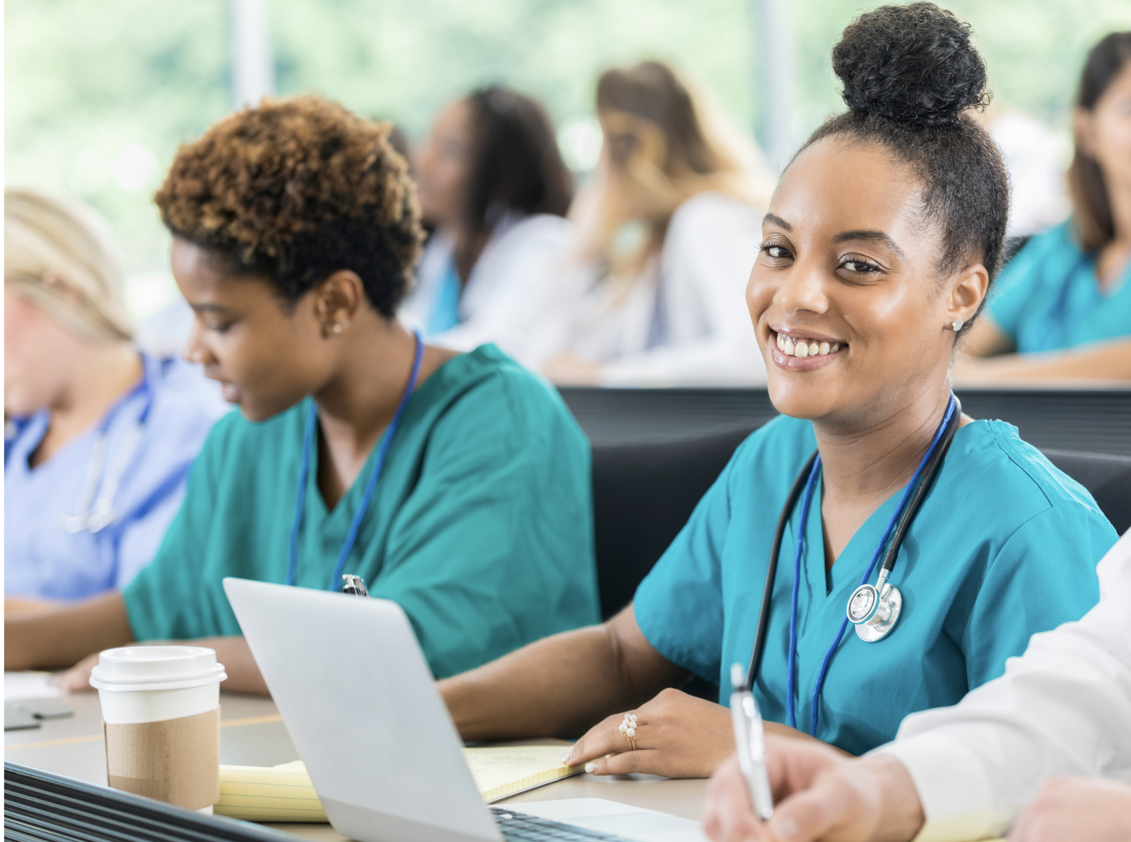 Smiling healthcare worker in teal scrubs sits at a desk with a laptop in a bright classroom or hospital setting, others at a table in the background